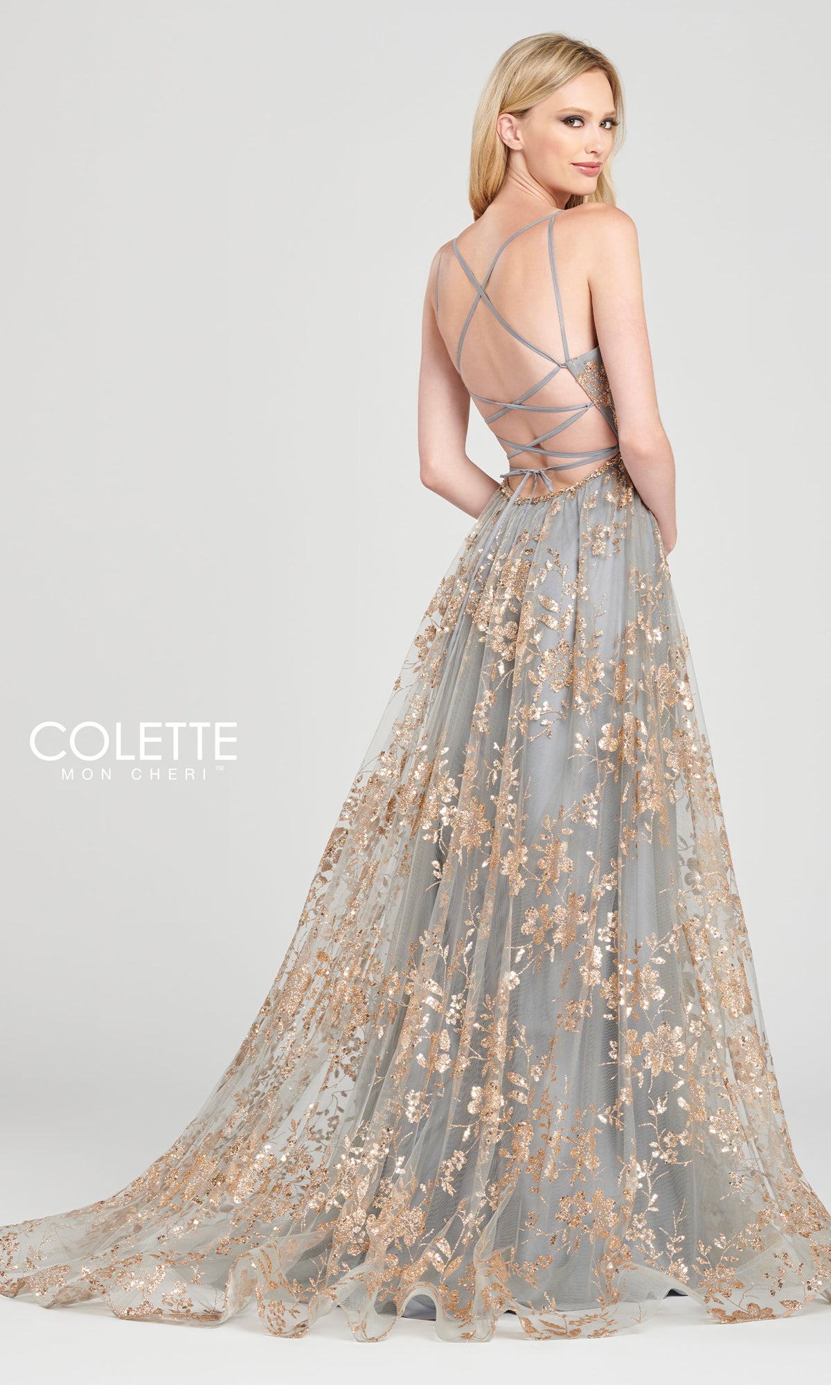 A woman stands slightly turned away in the Colette Gold and Pewter Long Prom Dress CL12006, featuring an a-line silhouette with gold floral embroidery and shimmery prints over a sheer gray skirt.